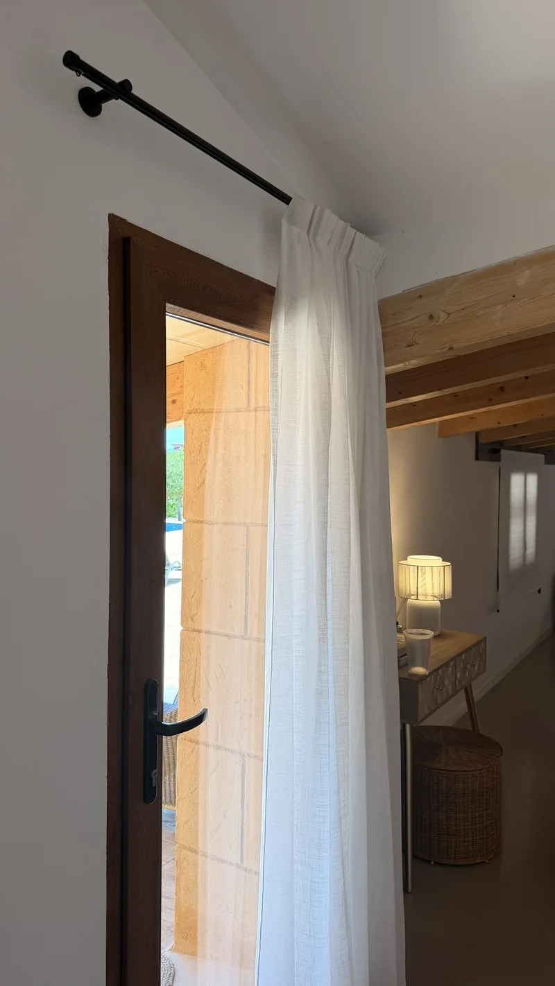 Barra negra con soporte a pared sobre una puerta de madera, con visillo blanco de lino