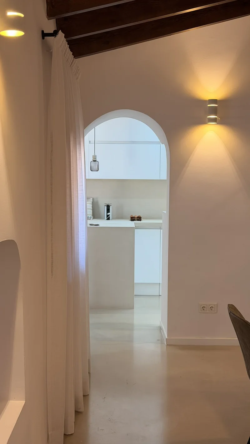 Paso bajo vigas de madera con visillo blanco de lino junto a un arco que da a la cocina