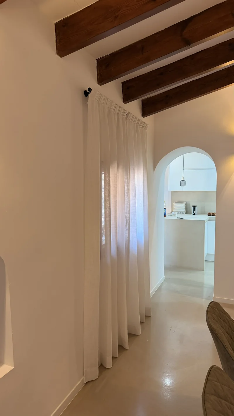 Visillos blancos de lino colgados de una barra negra bajo las vigas, con el arco de la cocina al fondo