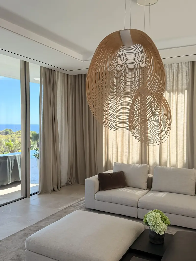 Salón con visillos y cortinas recogidas junto a los ventanales, con vistas al mar y una gran lámpara de fibras