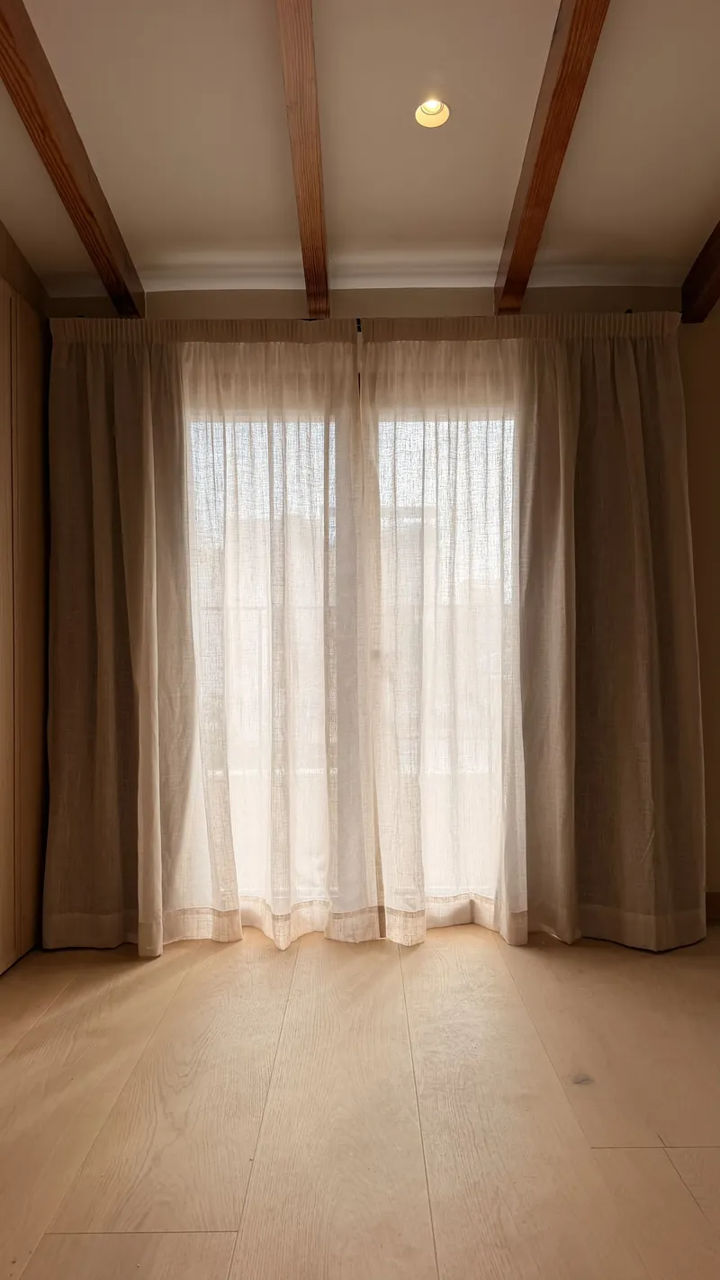 Dormitorio con vigas vistas, visillo de lino en mezcla y cortinas blackout a los lados de la ventana