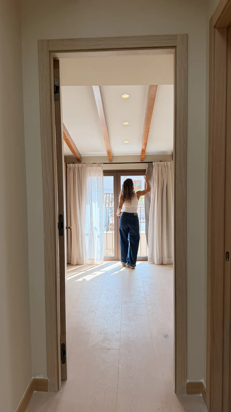 Vista desde la puerta de un dormitorio con vigas hacia un ventanal con visillo y luz natural