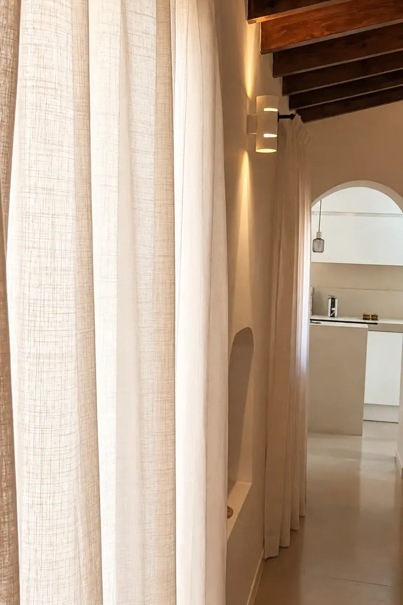 Visillo de lino blanco con caída natural junto a una pared con hornacina y techo de vigas de madera