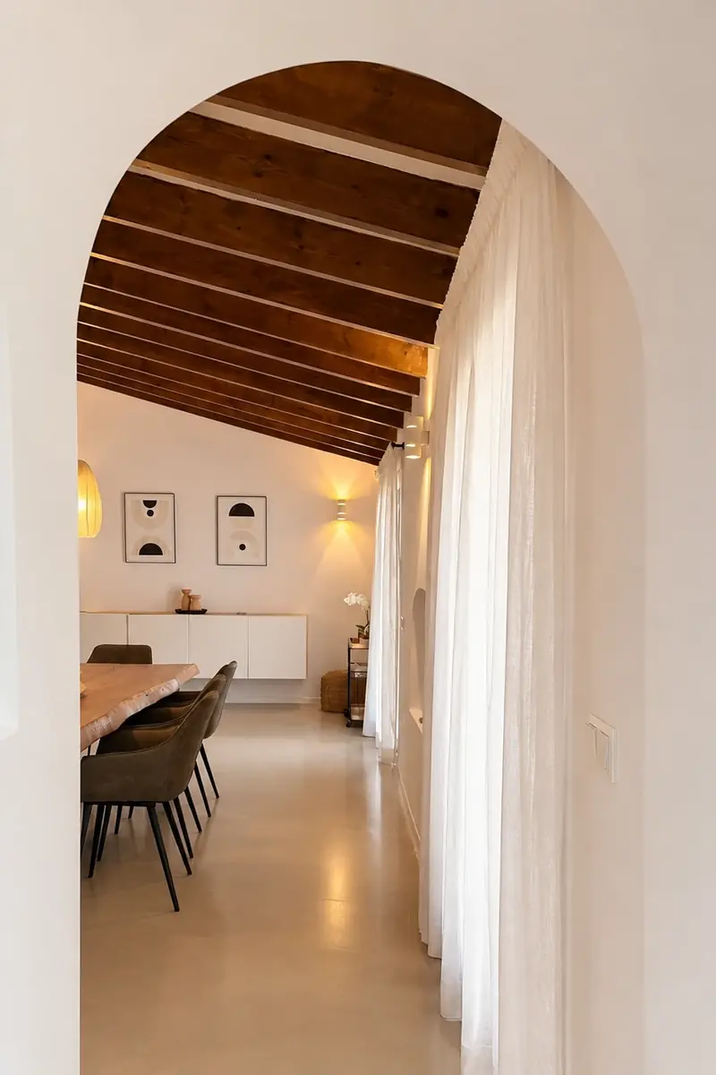 Visillos blancos de suelo a techo junto a un comedor con vigas de madera, vistos a través de un arco